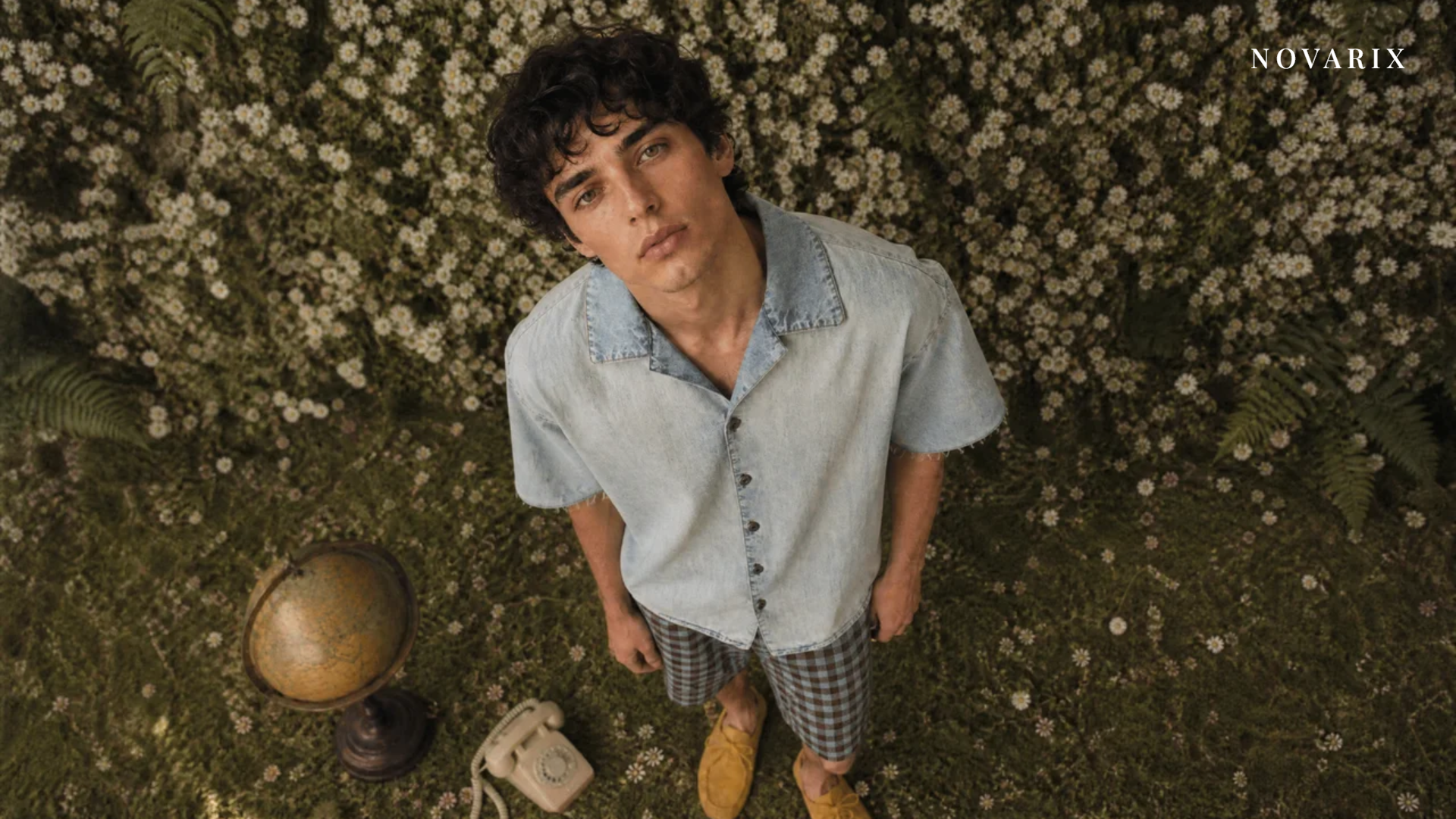 Young man in a denim shirt and gingham shorts standing in a field of daisies with a vintage globe and telephone
